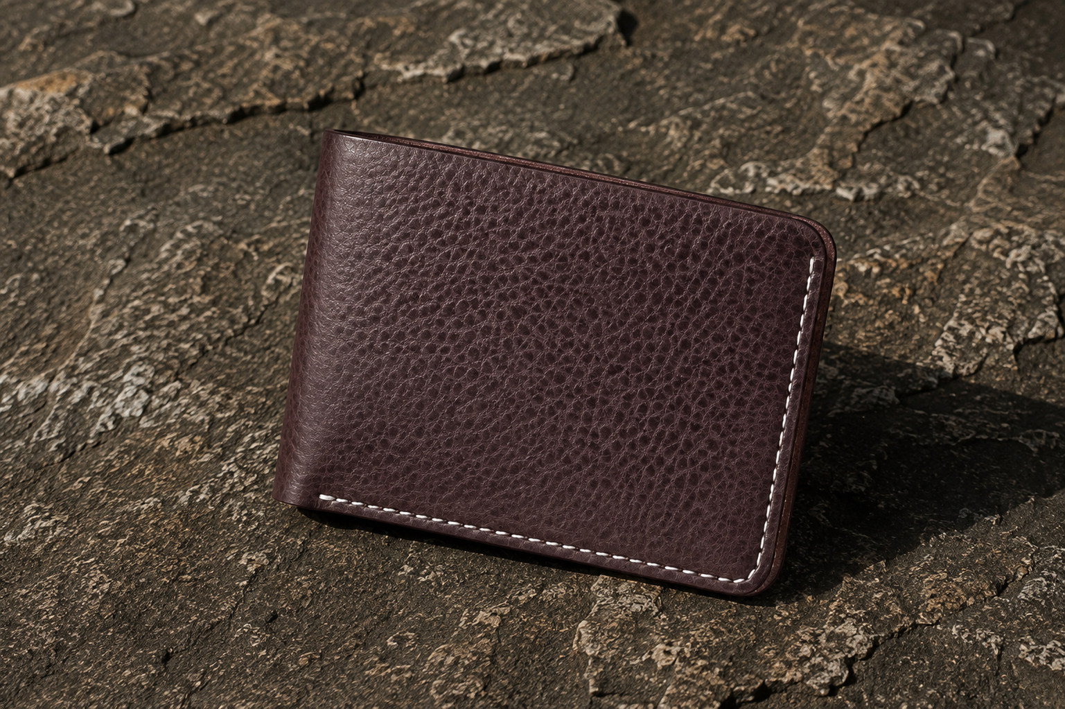 A dark brown pebbled leather bifold wallet with white contrast stitching, resting on a textured dark stone surface.