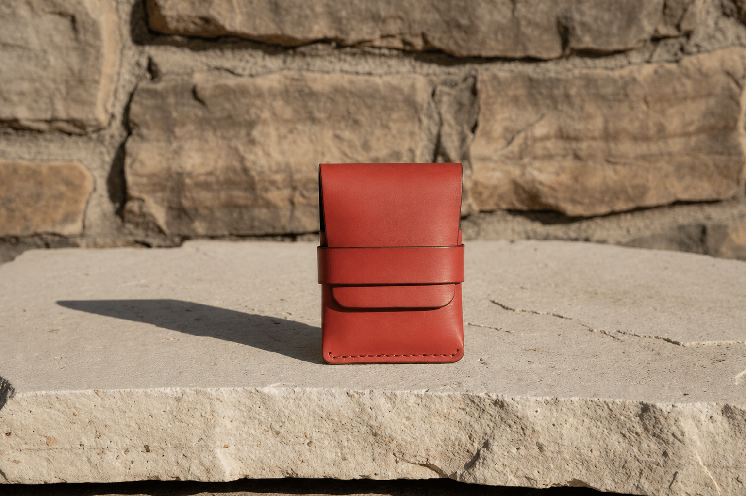 A small red leather card holder or wallet with a strap closure, standing upright on a stone surface in front of a stone wall.