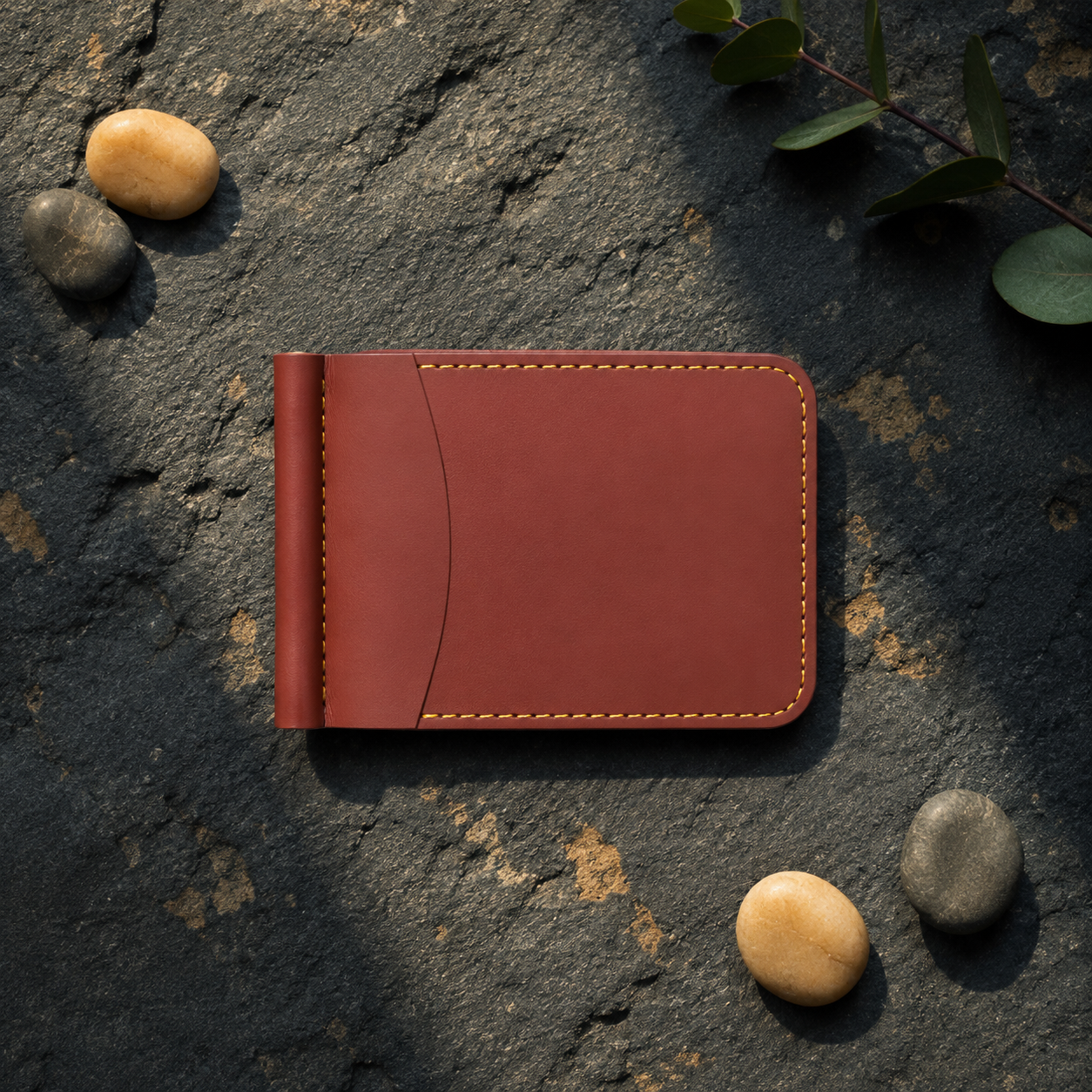 A reddish-brown leather money clip wallet with light tan stitching, styled on a dark textured stone surface with small decorative pebbles and green leaves.