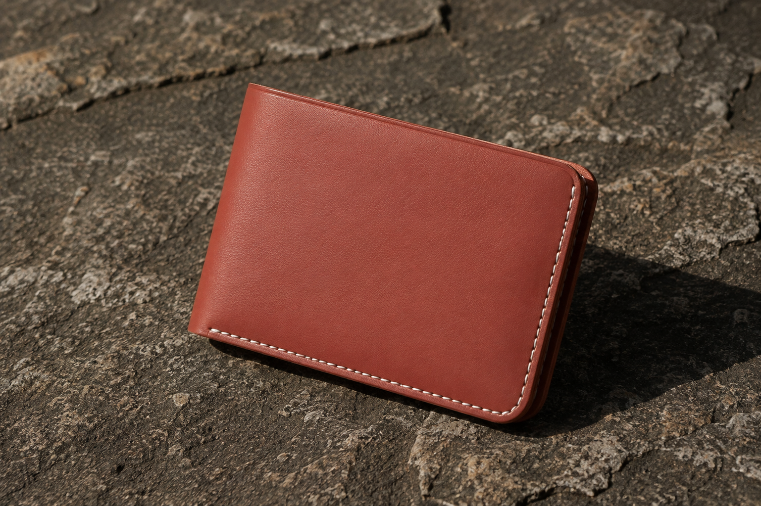 A reddish-brown leather bifold cardholder with white contrast stitching, resting on a textured dark gray stone surface.