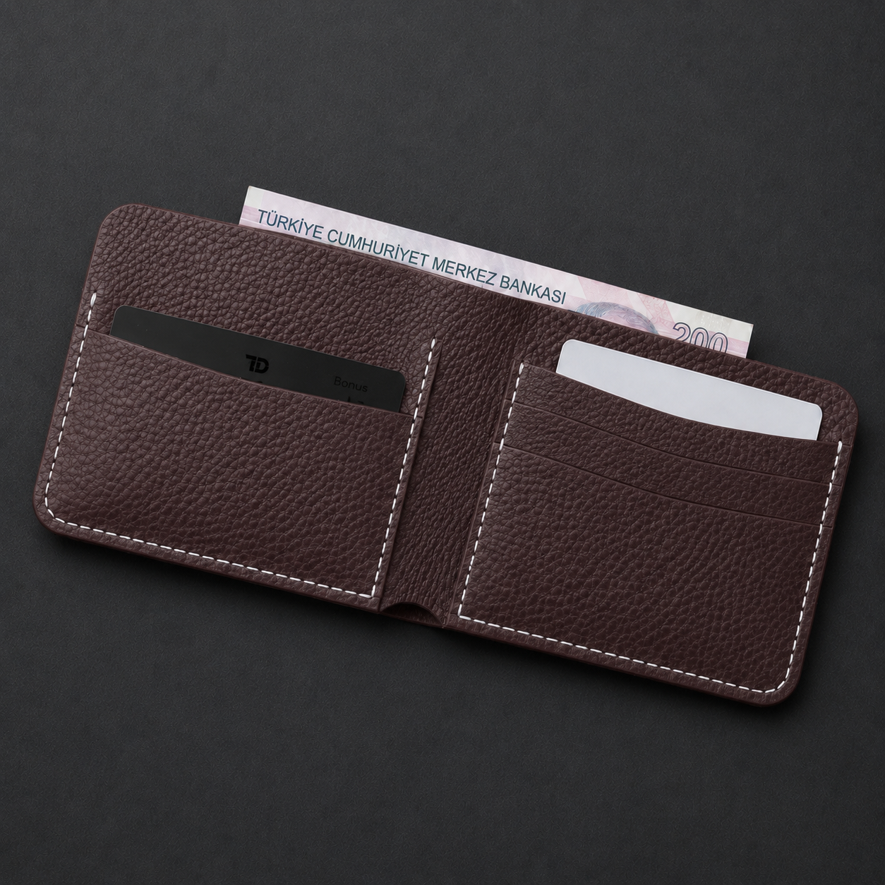 A dark brown pebbled leather bifold wallet with white contrast stitching, shown open on a dark gray background. The wallet contains card slots, a black ID card, and a Turkish 200 Lira banknote tucked behind it.
