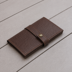 A dark brown textured leather card holder wallet with a snap button closure, resting on a light gray wooden plank surface.