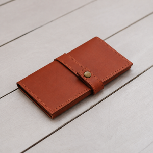 A brown leather wallet with a strap closure and brass snap button, resting on a light gray wooden plank surface.